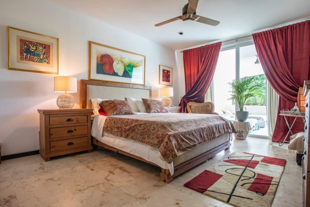 A bedroom in Bosque Los Aluxes Condo in Playa del Carmen features a large bed with patterned bedding, side tables with lamps, colorful artwork on white walls, and red curtains opening to a balcony with green plants.
