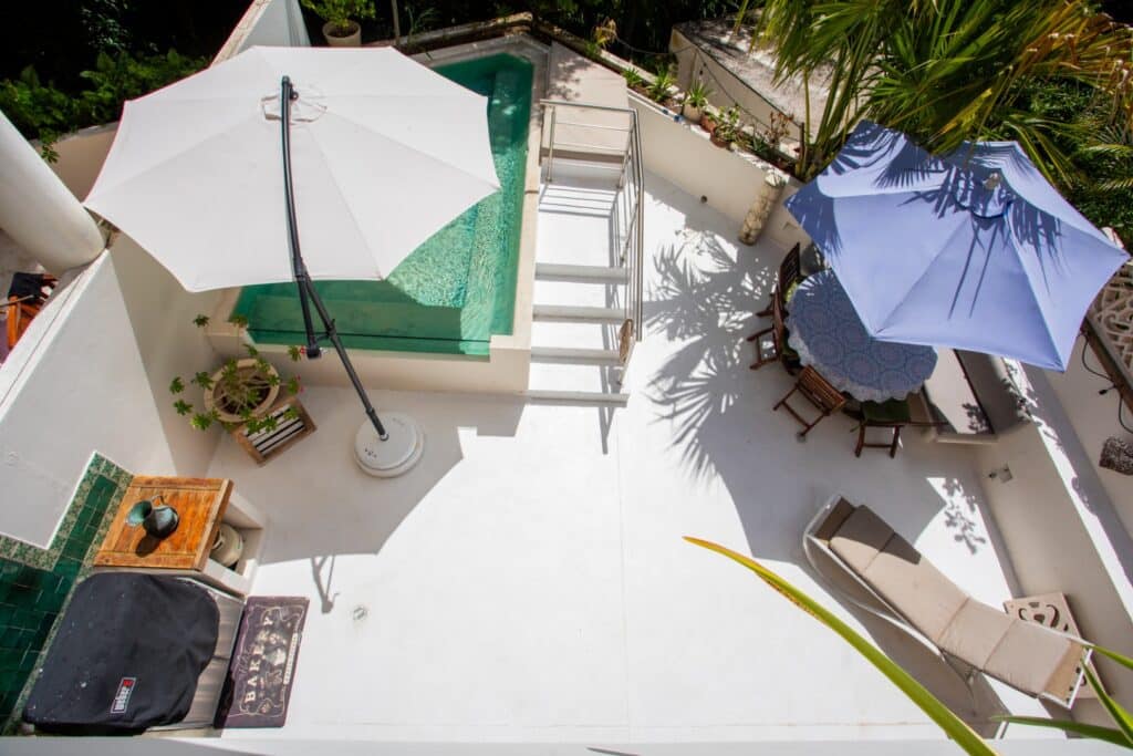 An outdoor terrace at Bosque Los Aluxes Condo in Playa del Carmen features a small pool, sun loungers, umbrellas, and potted plants, with shadows cast by surrounding palms.