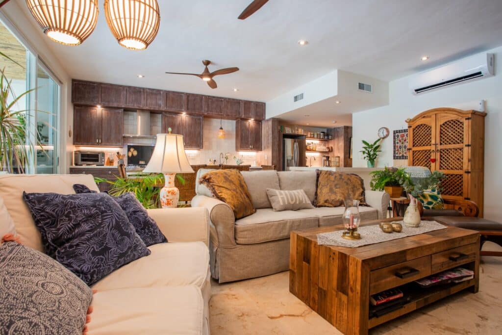 Living room with beige and white sofas, decorative pillows, wooden furniture, potted plants, and a kitchen area in the background at Bosque Los Aluxes Condo in Playa del Carmen.
