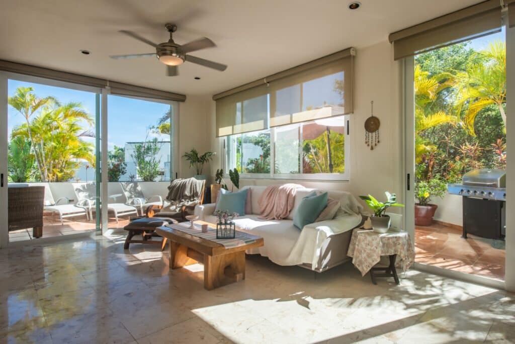 Living room with large windows, sliding glass doors opening to a terrace with tropical plants, a ceiling fan, and a white sofa with pastel cushions in Bosque Los Aluxes 301, Playa del Carmen.
