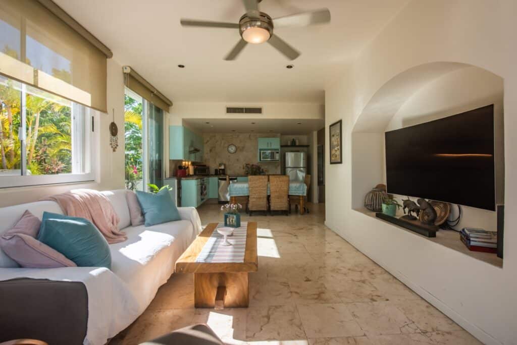 Interior view of the Bosque Los Aluxes 301 Penthouse in Playa del Carmen, featuring a living room with a large window, a ceiling fan, a TV, and an open kitchen with dining area.