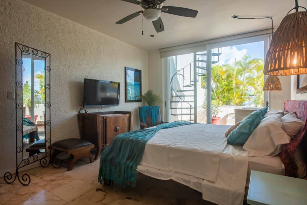 A bedroom in Bosque Los Aluxes 301 Penthouse in Playa del Carmen featuring a bed, a wall-mounted TV, a mirror, and a balcony with a spiral staircase, surrounded by lush greenery.