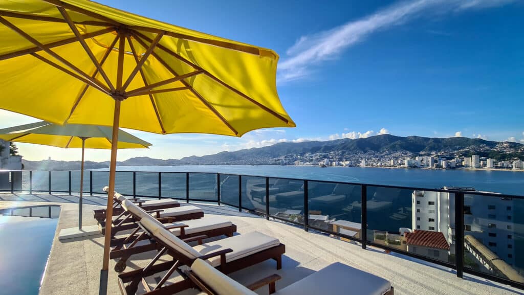 A rooftop terrace in Casa Buenos Aires, Acapulco, with lounge chairs under yellow umbrellas and a stunning view of the bay and city skyline against a blue sky.