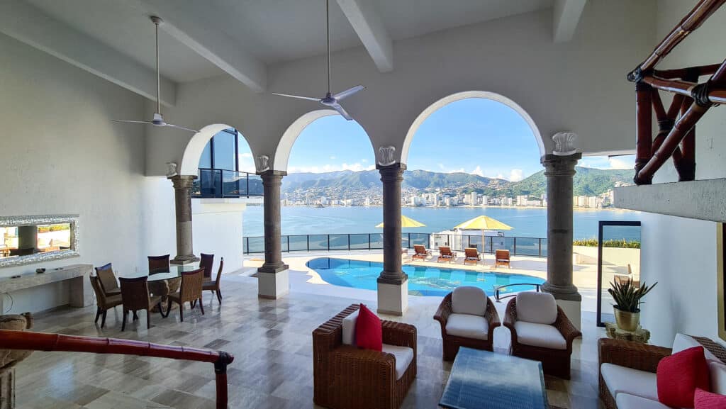 Interior of Casa Buenos Aires in Acapulco's Las Brisas with a view of the ocean, featuring a pool, arched windows, and a mix of wicker and wooden furniture.