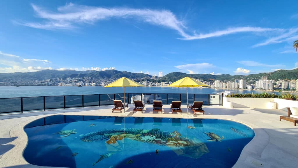 A rooftop pool with a view of Acapulco's skyline and mountains in the background, featuring four lounge chairs, yellow umbrellas, and a painted ocean scene on the pool floor.