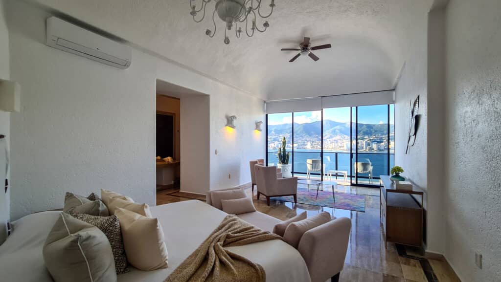 Living room with white textured walls, a chandelier, ceiling fan, large glass windows with ocean and mountain views, and modern furniture in Casa Buenos Aires, Acapulco.