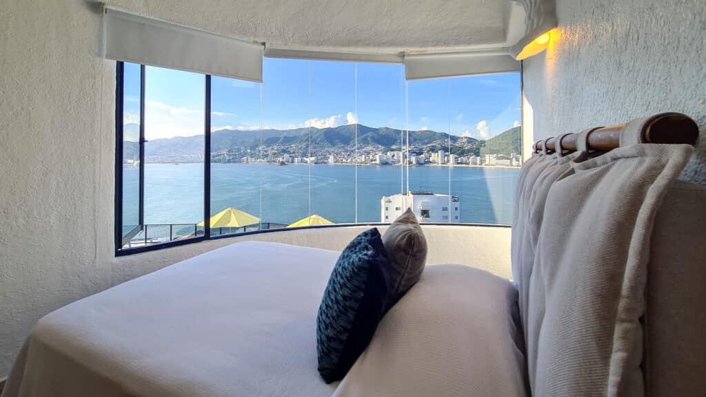 A bedroom in Casa Buenos Aires in Acapulco features a large window with a panoramic view of the bay, mountains, and city skyline, with a cozy bed and soft pillows.
