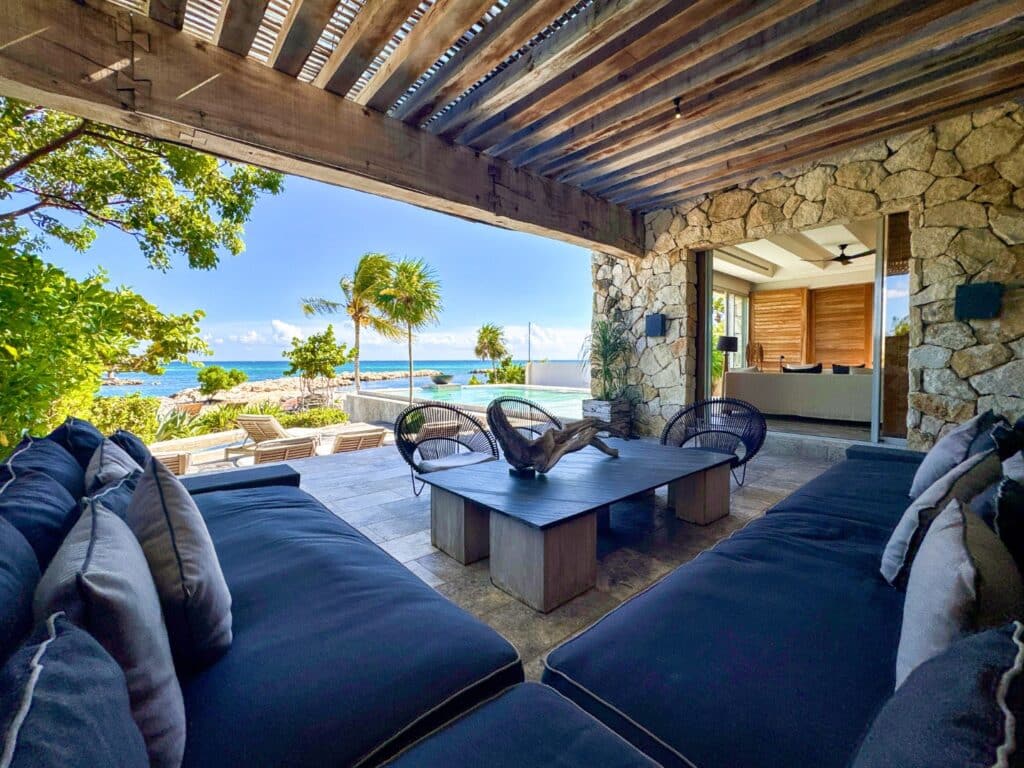 A shaded outdoor seating area with dark cushions overlooks a beachfront view of turquoise water, palm trees, and clear skies at Villa Caleta in Puerto Aventuras.