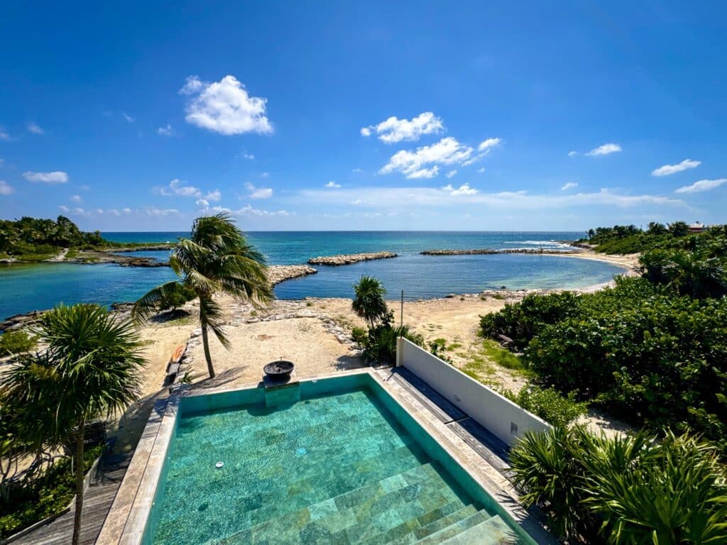 A view of the beachfront at Villa Caleta in Puerto Aventuras, featuring a private pool, lush greenery, and a sandy shoreline with the ocean and blue sky in the background.