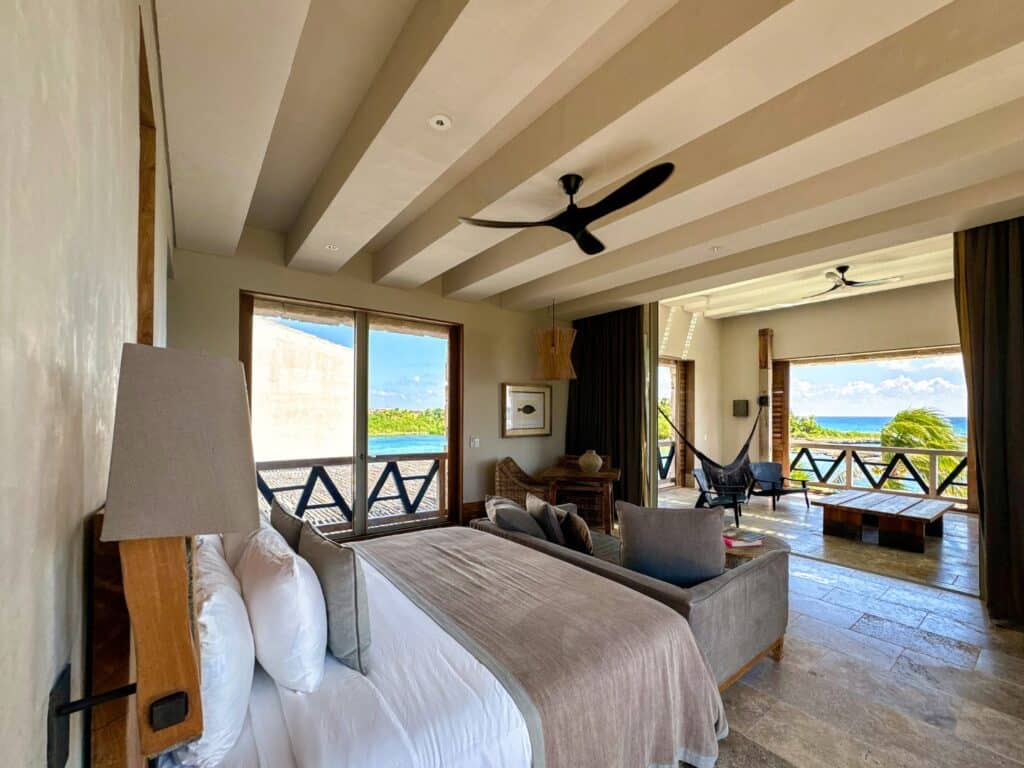 Interior view of Villa Caleta, Puerto Aventuras, showing a bedroom with large windows, wooden accents, balconies overlooking the beachfront, and tropical scenery.