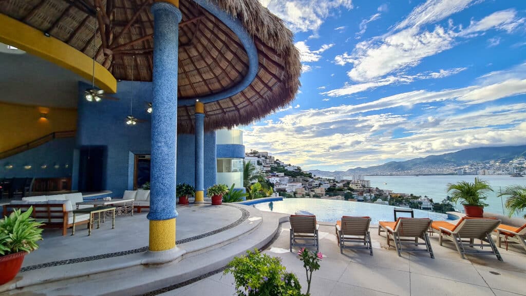 A view from Casa Carey in Acapulco, featuring a swimming pool with lounging chairs, colorful potted plants, and an expansive ocean and mountain backdrop under a partly cloudy sky.