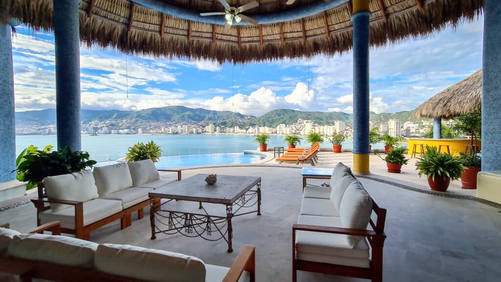 A spacious outdoor lounge area at Villa Carey in Acapulco features comfortable white seating, potted plants, an infinity pool, and a panoramic view of the city and mountains.