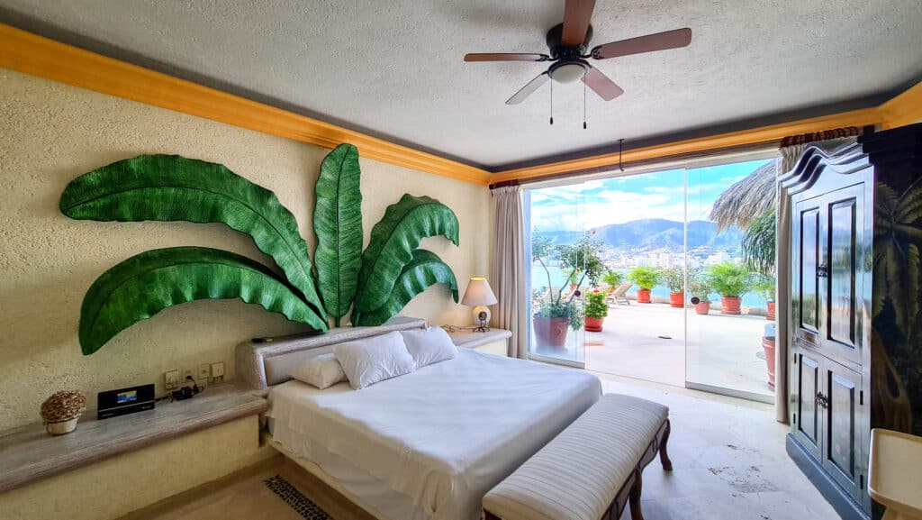 Bedroom in Villa Carey, Acapulco, featuring a tropical leaf wall art, a large sliding glass door opening to a terrace with mountain and city views.