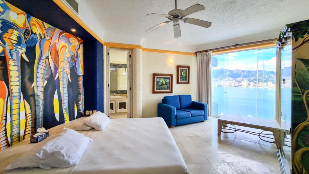A spacious bedroom in Villa Carey, Acapulco, with a large window offering a view of the water and mountains, featuring vibrant artwork on the headboard wall and a blue sofa.