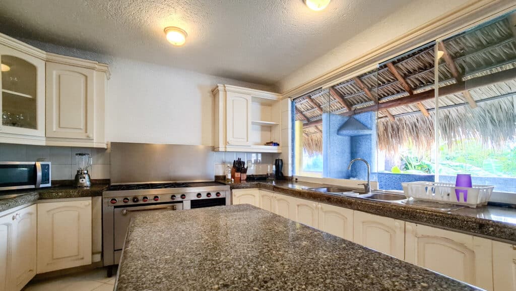 A spacious kitchen in Villa Carey in Acapulco features cream-colored cabinets, a granite countertop, and a large window overlooking a thatched roof patio.