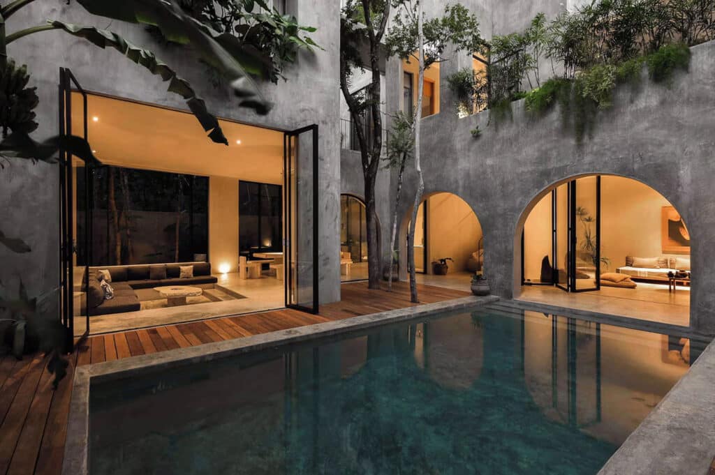A modern house at Casa Shalva in Tulum's Aldea Zama features large glass doors opening to a wooden deck with a pool, revealing a cozy living area inside with warm lighting.