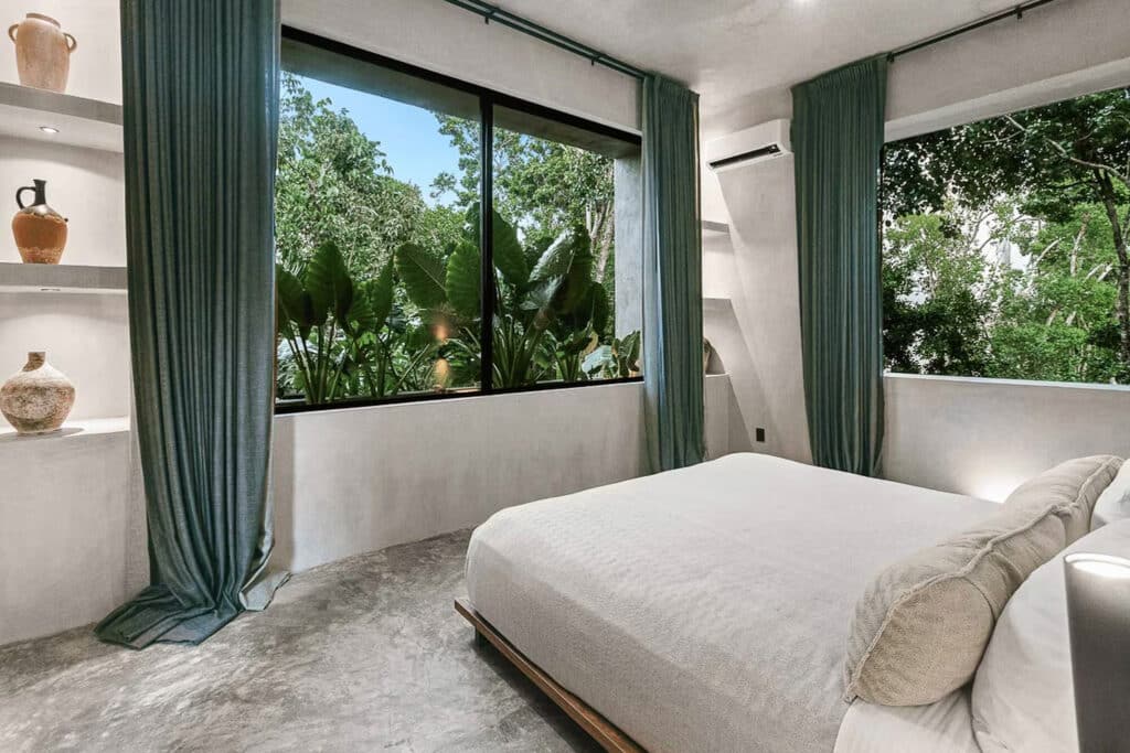 Interior view of Casa Shalva in Aldea Zama, Tulum, showing a bedroom with large windows, green curtains, and lush greenery outside.