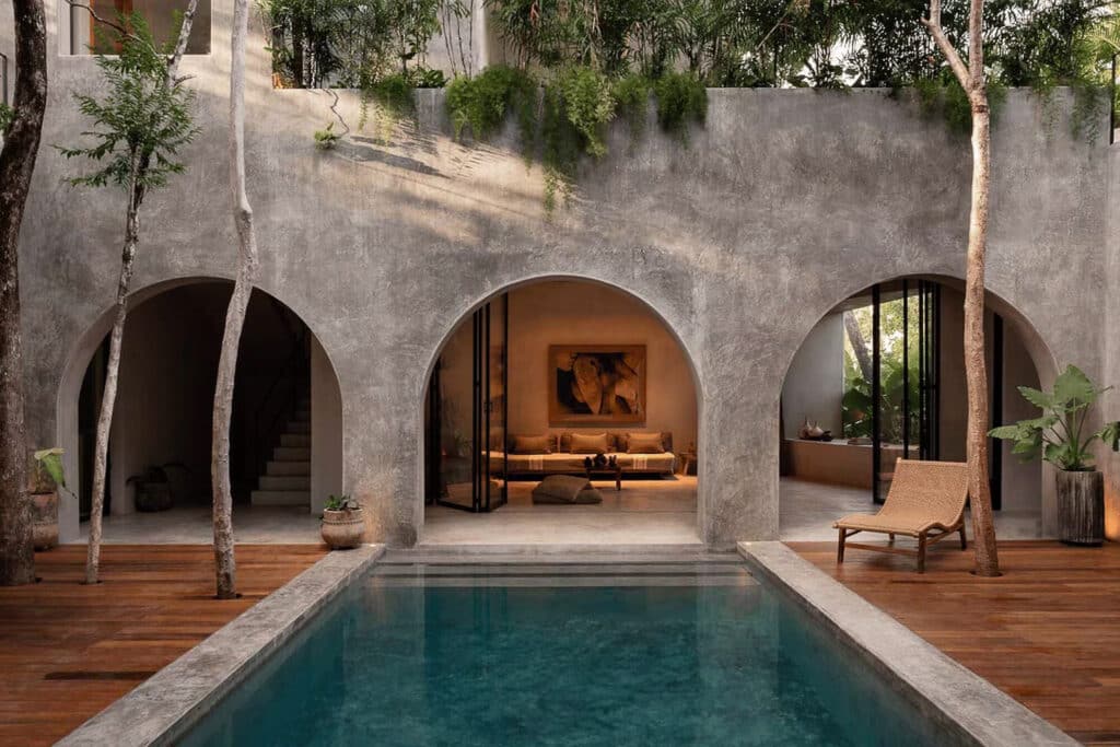 A modern house at Casa Shalva in Aldea Zama, Tulum, features an outdoor pool with arched openings leading to a cozy indoor seating area and lush greenery surrounding the space.