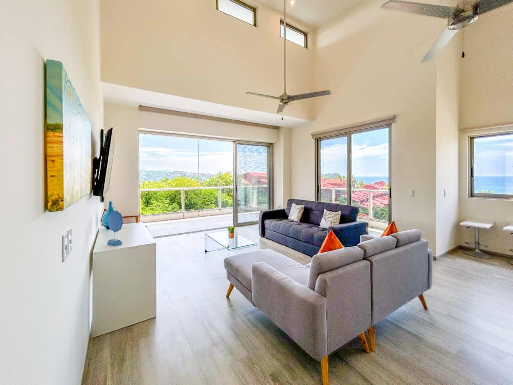 Living room in Cosmo PH5 beachfront residence in Huatulco, featuring large windows with ocean views, modern furniture, and a high vaulted ceiling with fans.