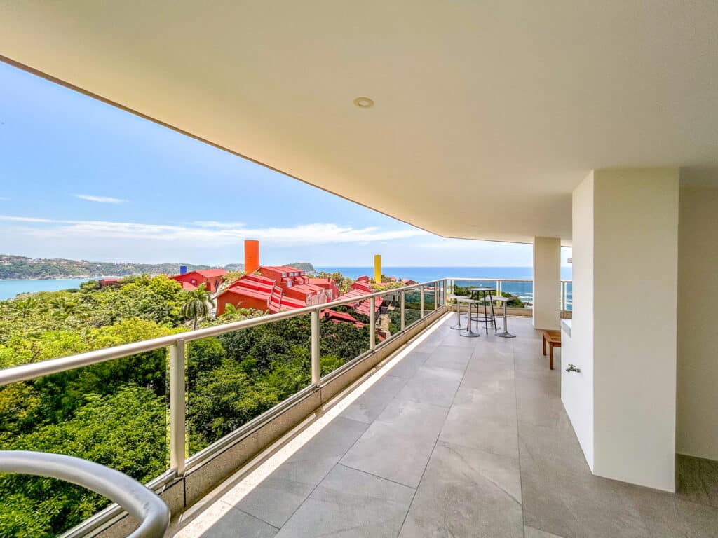 A spacious beachfront terrace at Cosmo PH5 in Huatulco features a view of lush greenery, colorful red-roofed buildings, and the ocean beyond on a clear day.