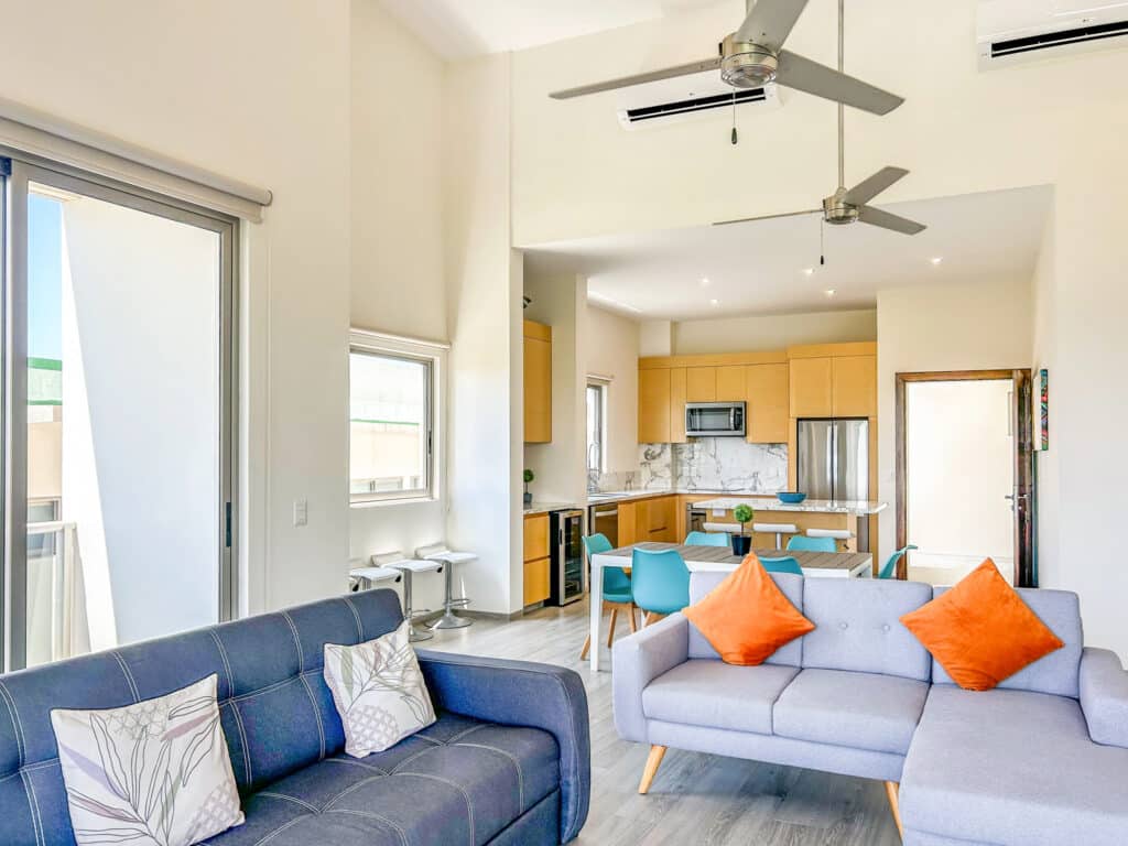 Living room and kitchen area of the Cosmo PH5 penthouse in Huatulco, featuring a modern open design, two sofas with colorful pillows, and large windows overlooking the beachfront.