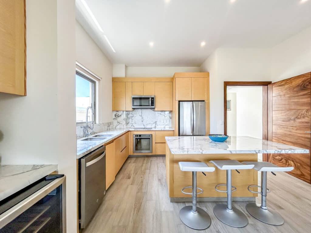 The kitchen in Cosmo PH5 features wooden cabinets, marble countertops, a stainless steel refrigerator, microwave, oven, and a marble-topped island with three white barstools,.