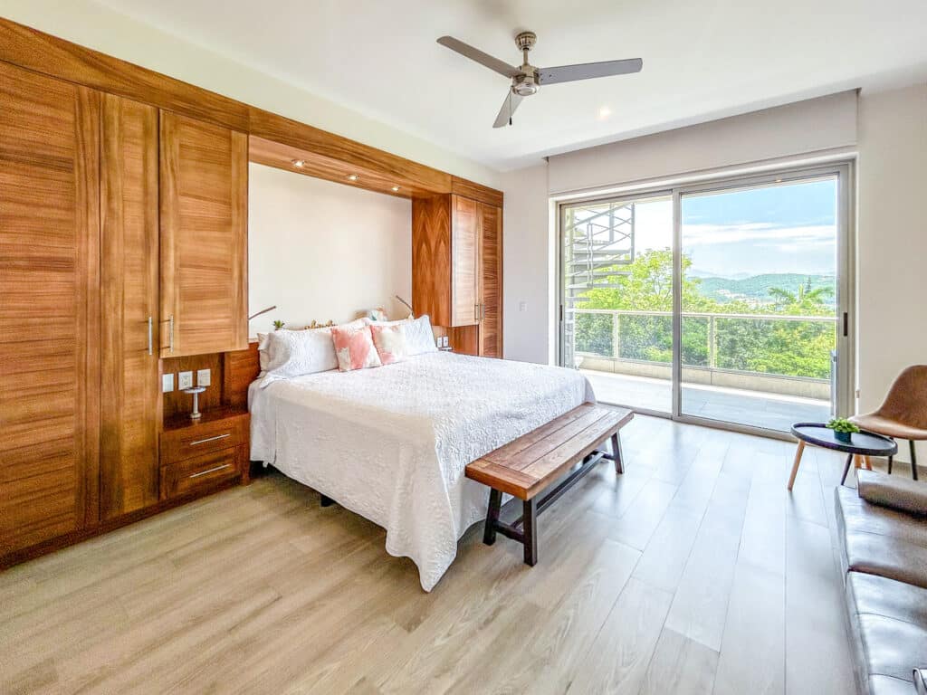 Bedroom interior of Cosmo PH5 in Huatulco with a wooden built-in wardrobe, a large bed with white bedding, a sliding glass door leading to a balcony with views of greenery and the sky.