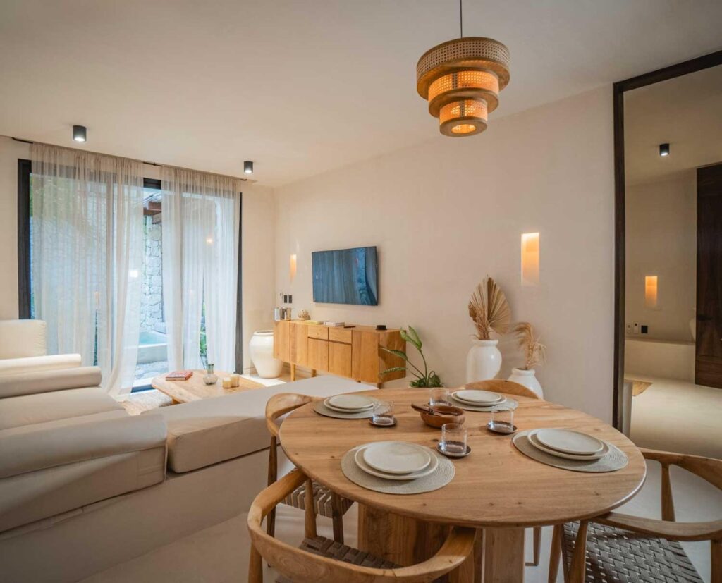 Living area of a Namas 13 residence in La Veleta, Tulum, featuring a round wooden dining table set with plates and glasses, a white sofa, and a wooden media console under a wall-mounted TV.