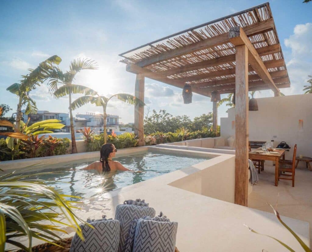 A woman relaxes in a rooftop pool at Namas 13 in La Veleta, Tulum, with a wooden pergola, lush tropical plants, and a clear blue sky overhead.