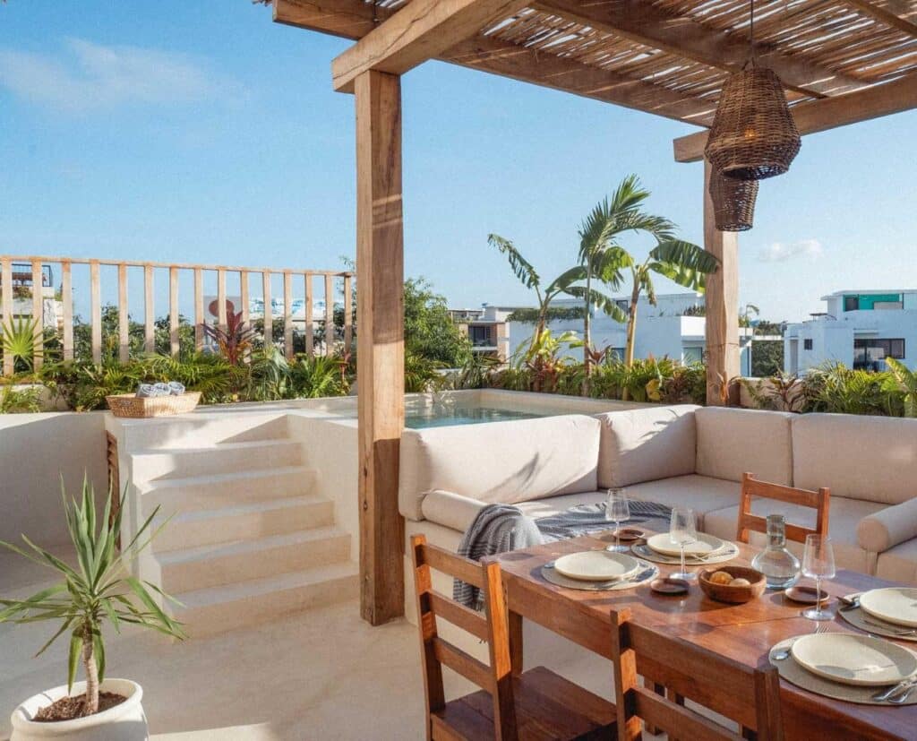 An outdoor rooftop patio in La Veleta, Tulum, featuring a dining table set and a cozy seating area under a wooden pergola overlooking lush greenery and neighboring modern buildings.