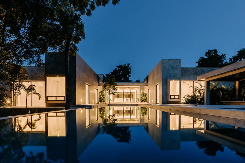 Modern Villa Amanai in Tulum features sleek architecture, illuminated interiors, and a reflective pool with lush greenery in the Rancho Viejo area.