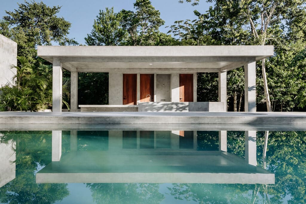 A modern concrete house with three wooden doors, situated by a reflecting pool surrounded by lush green trees in Villa Amanai, Tulum.
