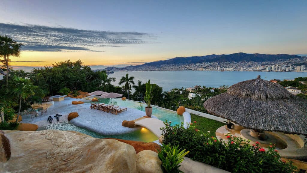 A view of Villa Gitana in Acapulco showing a pool area with lounge chairs, umbrellas, lush greenery, and a thatched hut overlooking the city skyline and bay at sunset.