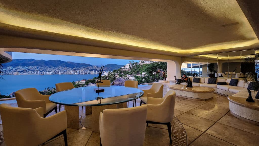 An elegant living area in Villa Gitana, Acapulco, with large windows offering views of the bay and mountains, featuring a round dining table and a spacious seating area.