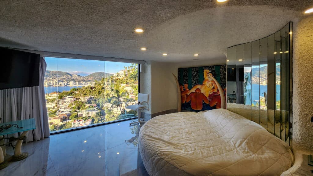 A bedroom in Villa Gitana, Acapulco, features a large window with a view of the city and mountains, a circular bed, abstract artwork, and mirrored closet doors.