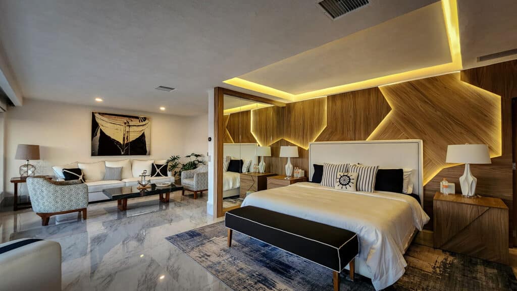 Interior view of a spacious bedroom in Villa Gitana, Acapulco, featuring a white bed with black and striped pillows, modern wooden accent wall with LED lighting, and adjacent sitting area.