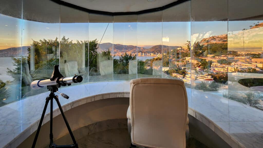 A telescope looking out from Villa Gitana in Acapulco, offering panoramic views of the city, mountains, and coastline through curved glass windows.