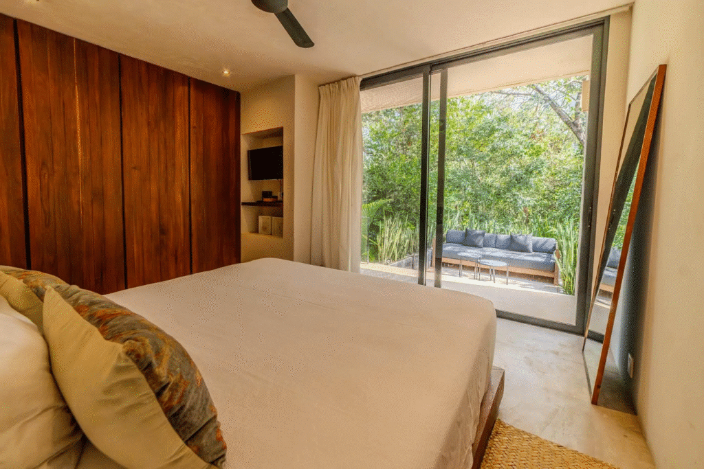 A bedroom at Villa Kaan in Tulum features a large bed with floral pillows, a wooden accent wall, and a sliding glass door opening to a balcony with outdoor seating and lush greenery.