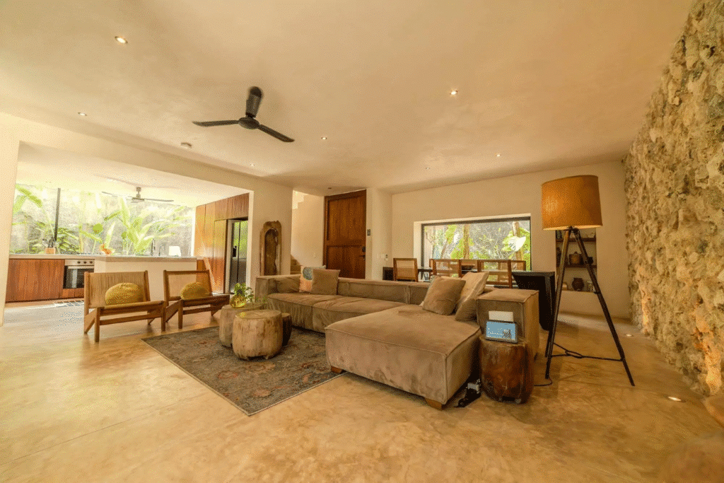 Living room in Villa Kaan, Tulum, with a beige sectional sofa, wooden chairs, a stone accent wall, and large windows overlooking greenery.