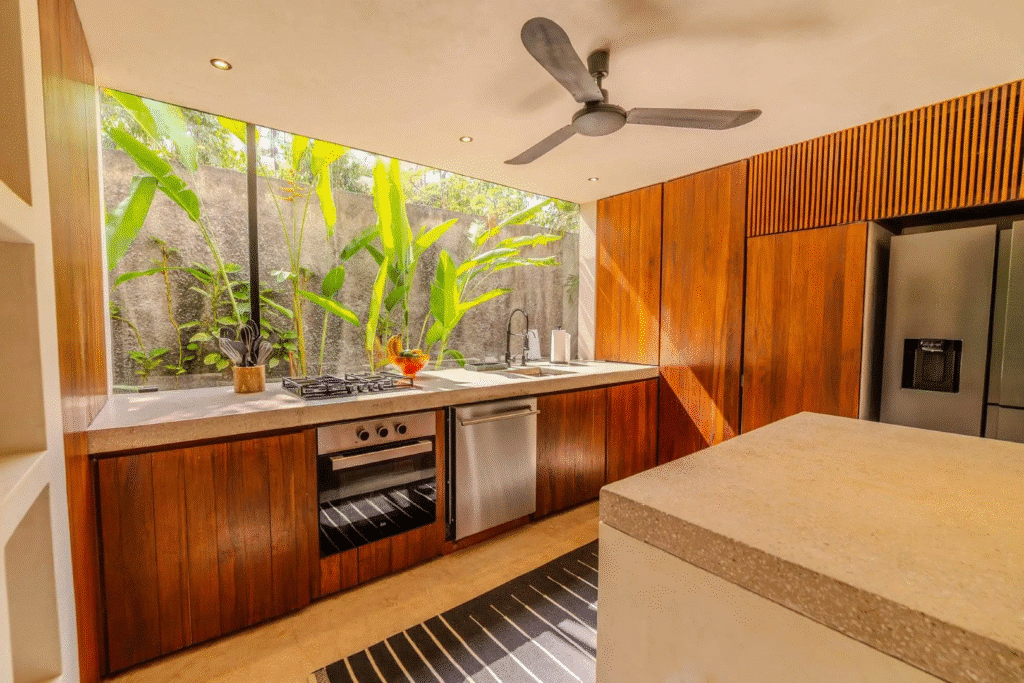 A modern kitchen at Villa Kaan in Holistika, Tulum, featuring a large window with lush greenery outside, wooden cabinets, a built-in oven, stainless steel appliances, and a ceiling fan.