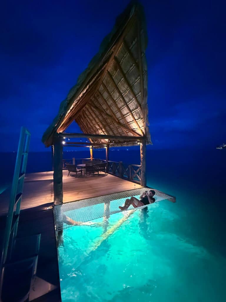 A person relaxing in a netted lounge by a lit swimming pool with a thatched roof structure, overlooking the ocean at Villa Tortuga in Cancun's beachfront area during nighttime.