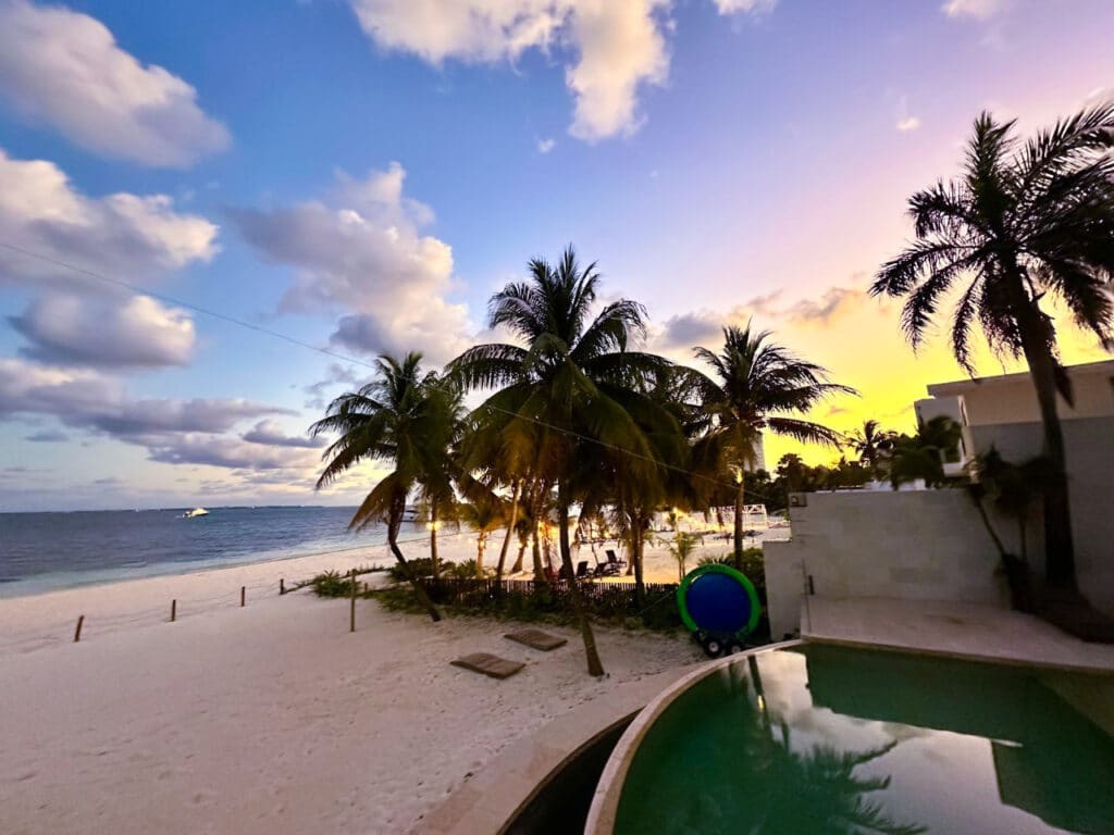 Beautiful beachfront view at Villa Tortuga in Cancun, featuring palm trees, a swimming pool, and a stunning sunset over the ocean.
