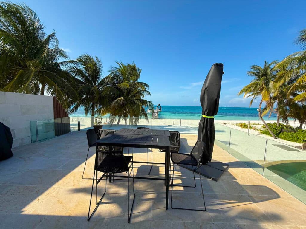 A beachfront terrace at Villa Tortuga in Cancun features a dining table with chairs, surrounded by palm trees, with the ocean view and clear blue sky in the background.