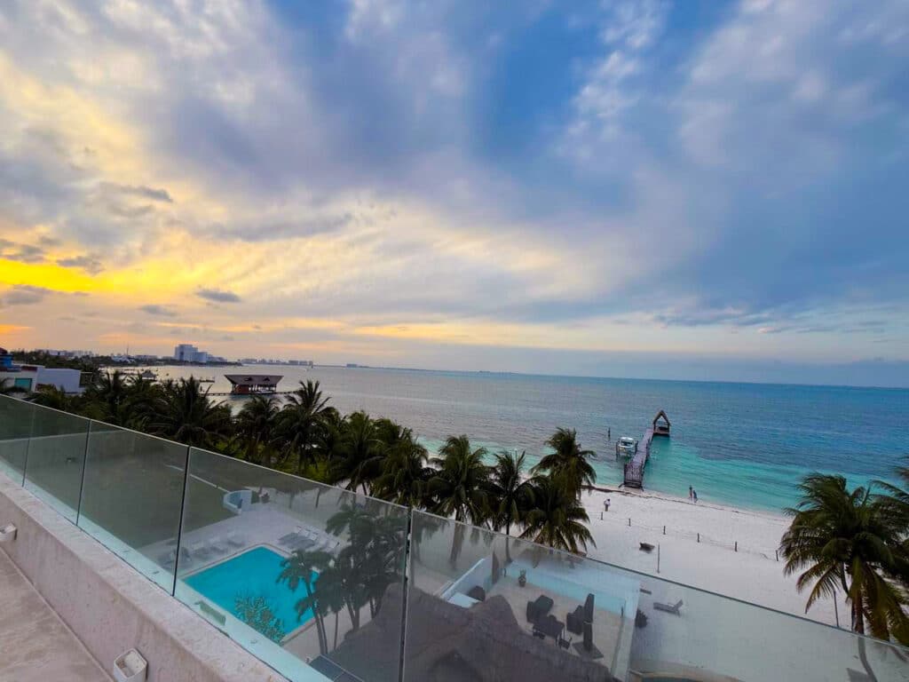 View of the beachfront in Cancun from Villa Tortuga, showing a swimming pool, palm trees, a pier extending into the ocean, and a colorful sky at sunset.