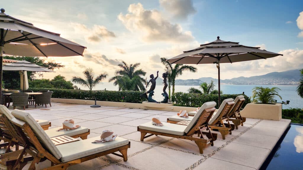 A spacious terrace at Villa Del Mar in Acapulco features sun loungers with towels, large umbrellas, elegant statues, tropical plants, and a view of the bay and mountains.