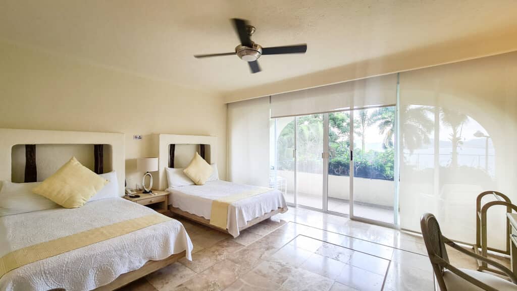 A bedroom in Villa Del Mar, Acapulco, featuring with white linens and yellow accents, a sliding glass door leading to a balcony with ocean views, and a ceiling fan.