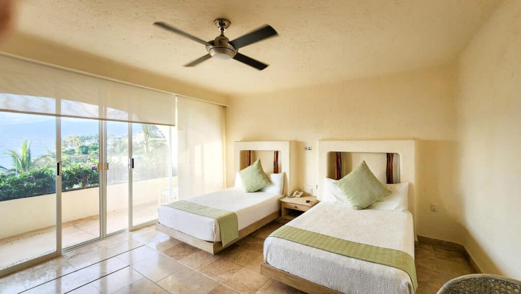 A bedroom at Villa Del Mar in Acapulco, featuring with green pillows, a ceiling fan, and a large sliding door opening to a balcony with ocean views.