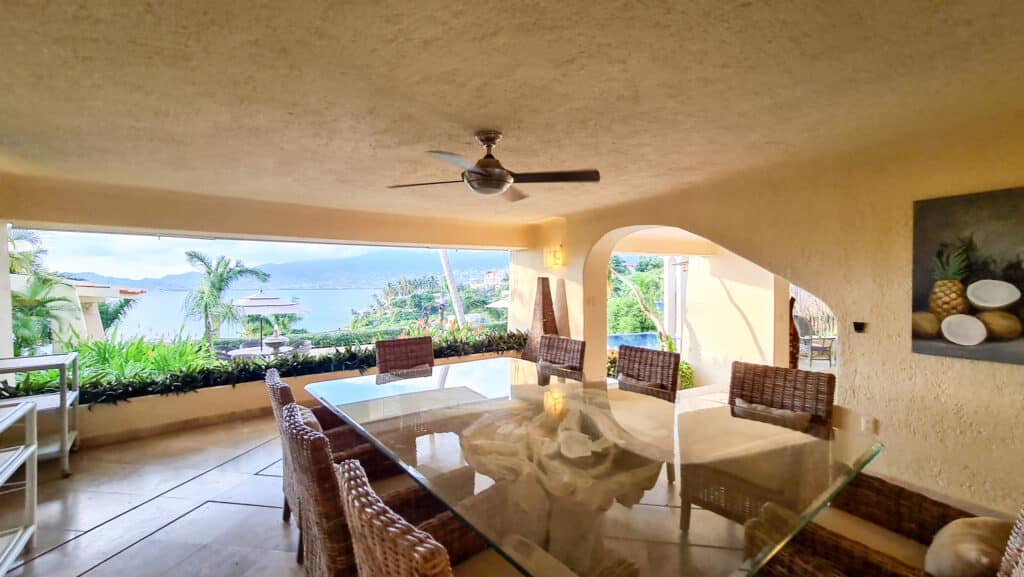 A dining area in Villa Del Mar, Acapulco, showcasing a glass-top table with wicker chairs, a ceiling fan, and a view of lush greenery, the pool, and the bay beyond.