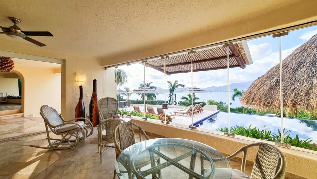 A view from inside a house at Villa Del Mar in Las Brisas, Acapulco, showing a glass-topped dining table, wicker chairs, and a large window that overlooks a pool and tropical landscape with mountains in the distance.