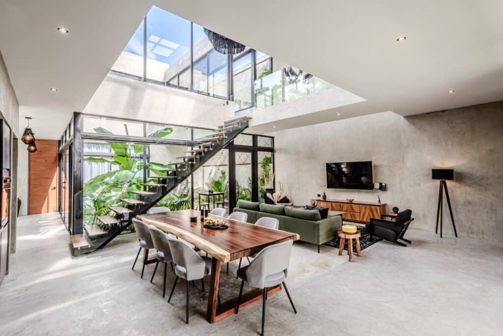 Interior view of Villa Karmond in Tulum, showing a spacious, modern living and dining area with large windows, a staircase, and lush greenery visible through the glass.